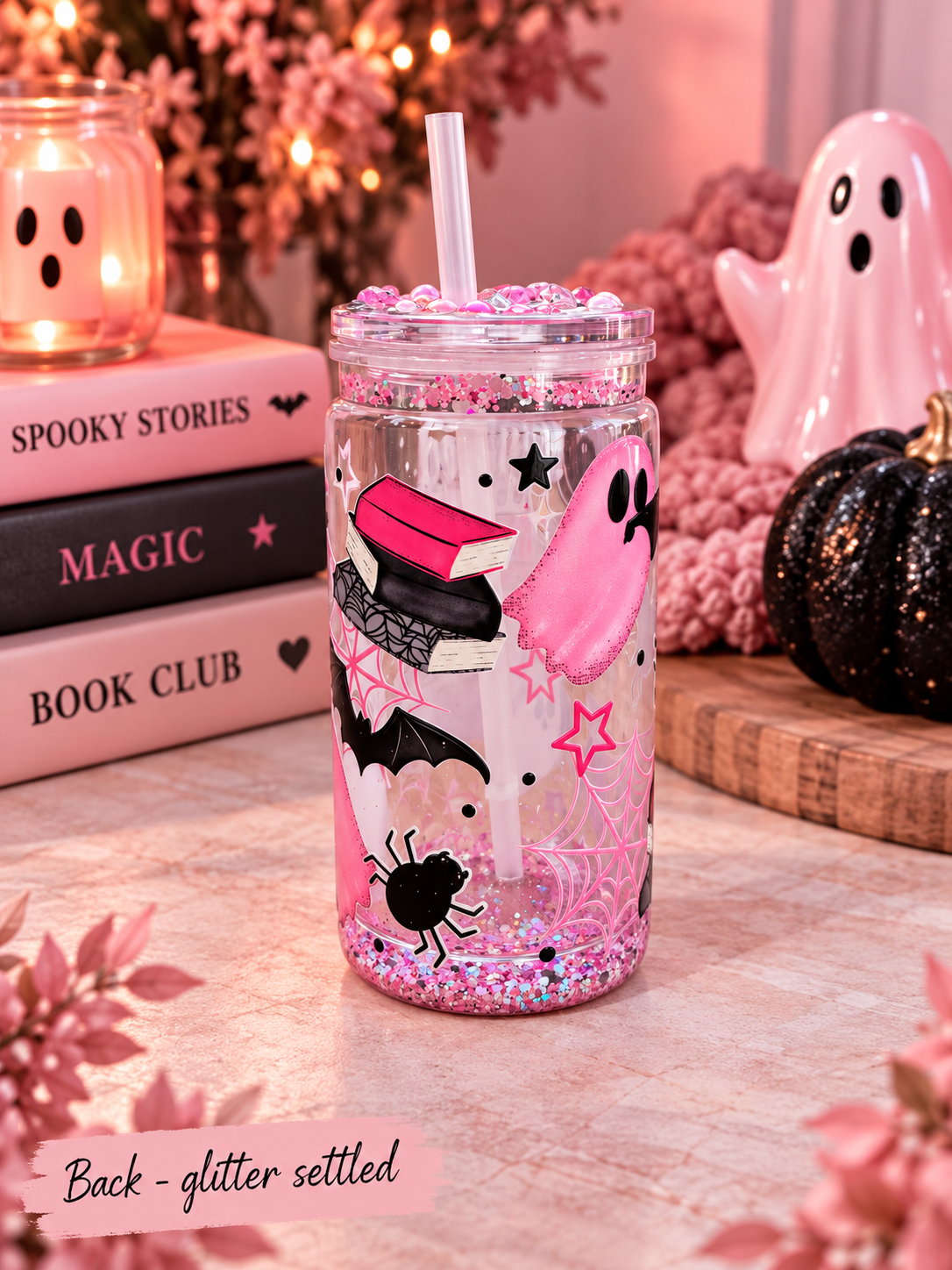 A clear plastic tumbler with a straw, decorated with pink glitter, a pink ghost, black spider, bat, and book illustrations, set on a tabletop with a cozy Halloween-themed background.