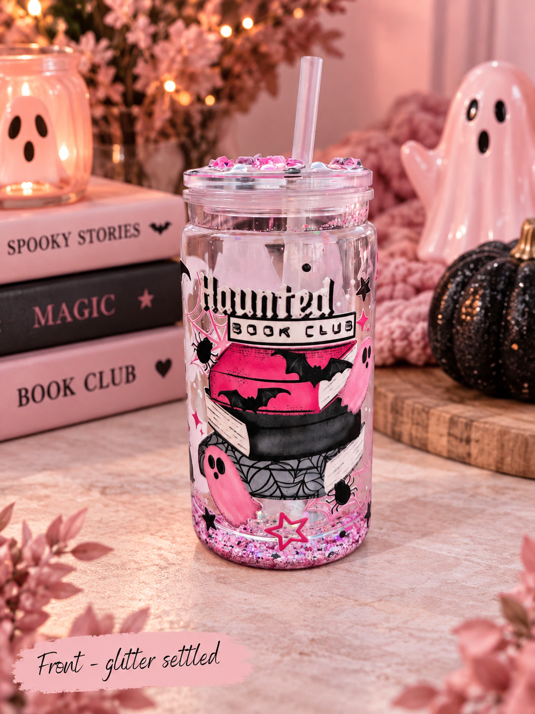 A clear glitter tumbler with a straw and rhinestone rim, decorated with a pink and black 'Haunted Book Club' book stack design, set on a tabletop with books, a ghost figurine, and warm string lights in the background.
