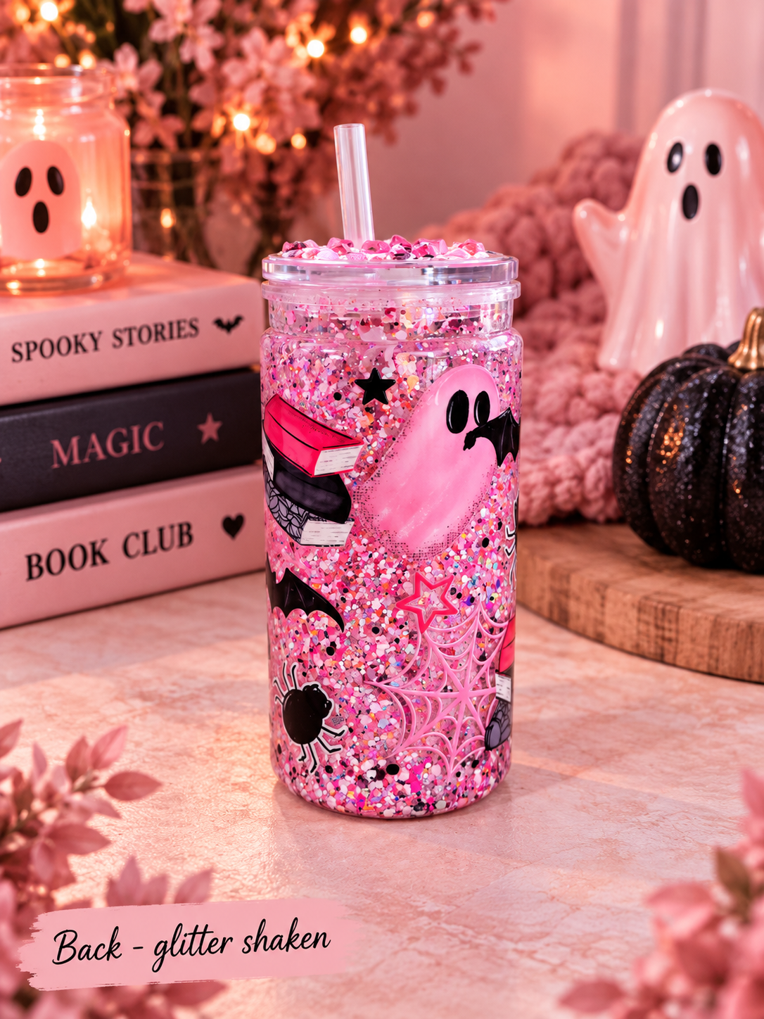 A pink glitter-filled tumbler with a clear lid and straw, decorated with a pink ghost, black spider, and stars, set on a tabletop with a candle, books, and a ghost figurine in the background.