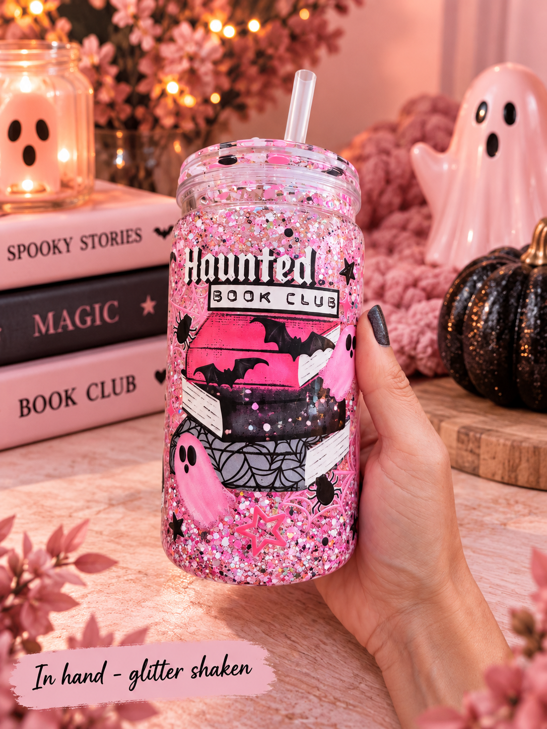 A hand holds a glittery pink mason jar tumbler with a straw, decorated with illustrations of stacked books, ghosts, and the text 'Haunted Book Club'. The background is a cozy, warm-toned autumn-themed setting with books, a candle, and a ghost figurine.