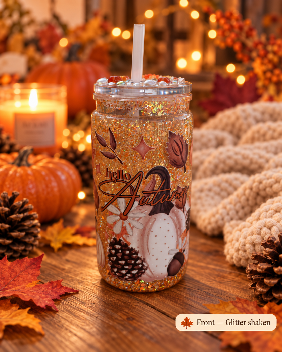A glitter-filled tumbler with a straw sits on a wooden table surrounded by autumn decor, including pumpkins, pinecones, fall leaves, and a knitted blanket. The tumbler has a clear lid with a white straw and multicolor fall-themed graphics including the words 'hello autumn.