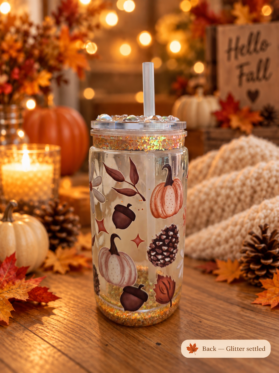 A clear mason jar tumbler with a lid and straw, decorated with autumn pumpkins, acorns, pinecones, and leaves, sitting on a wooden table surrounded by fall decorations including pumpkins, candles, and a knitted blanket.