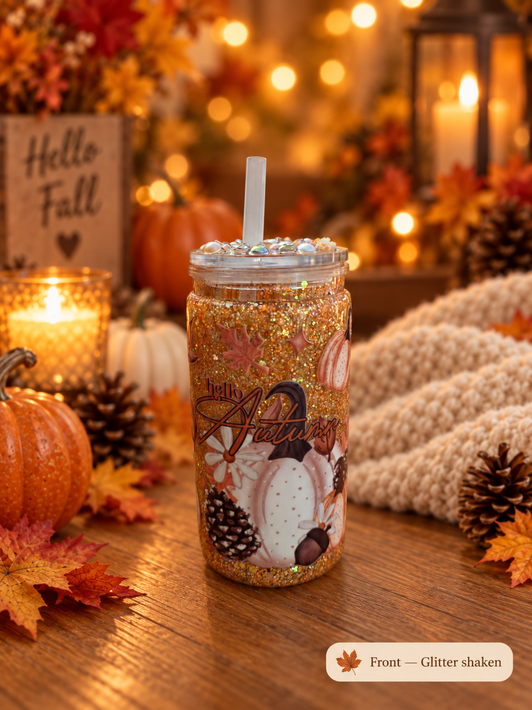 A glitter-filled clear tumbler with a straw sits on a wooden table in an autumn-themed setting with pumpkins, fall leaves, candles, and a knitted blanket. The tumbler features a printed design with pumpkins and the words 'Hello Autumn'.