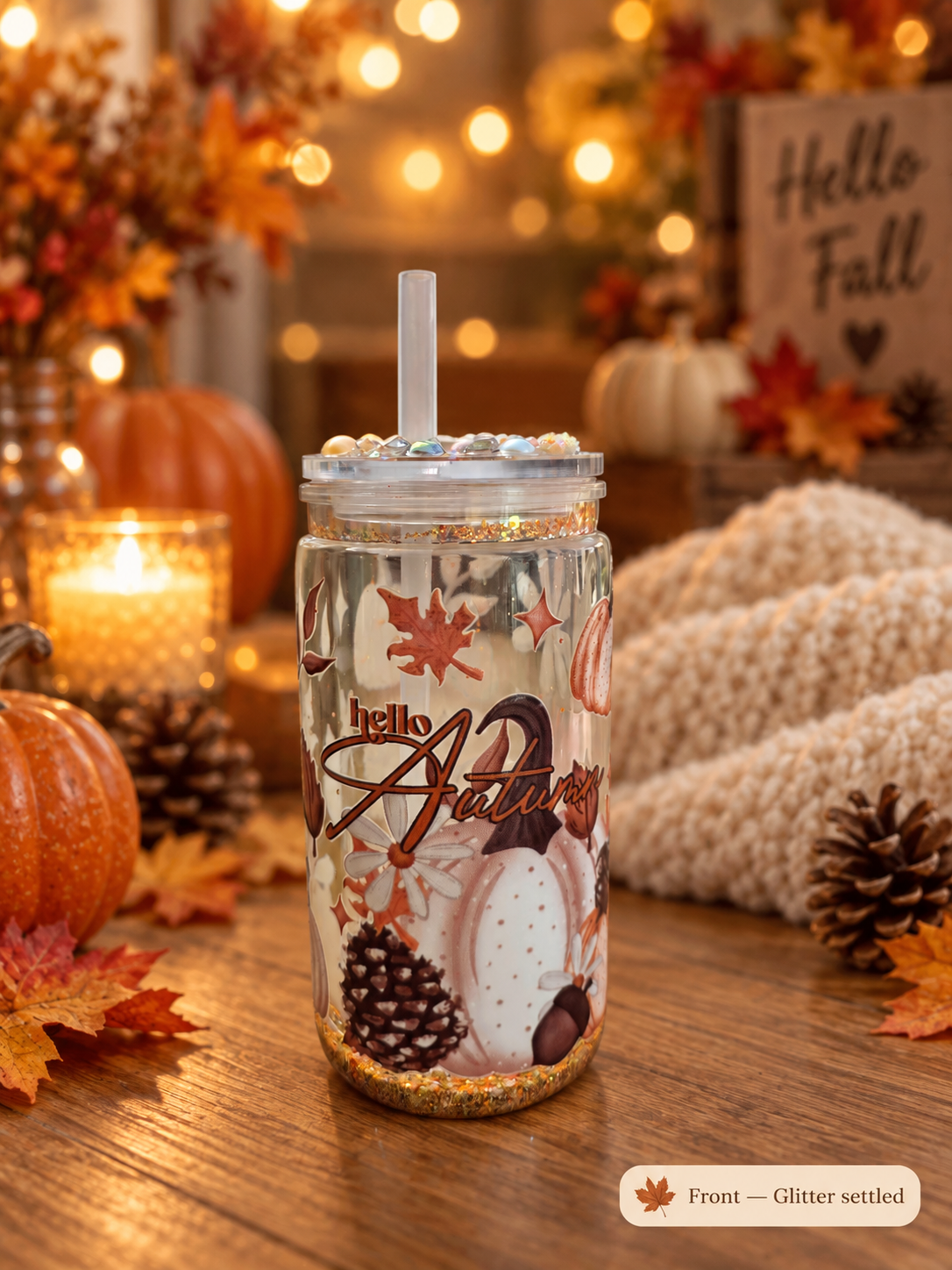 A clear glitter tumbler with a clear lid and straw, decorated with autumn illustrations including maple leaves, pumpkins, pinecones, and the words 'hello Autumn,' set on a wooden table with fall decorations in the background.