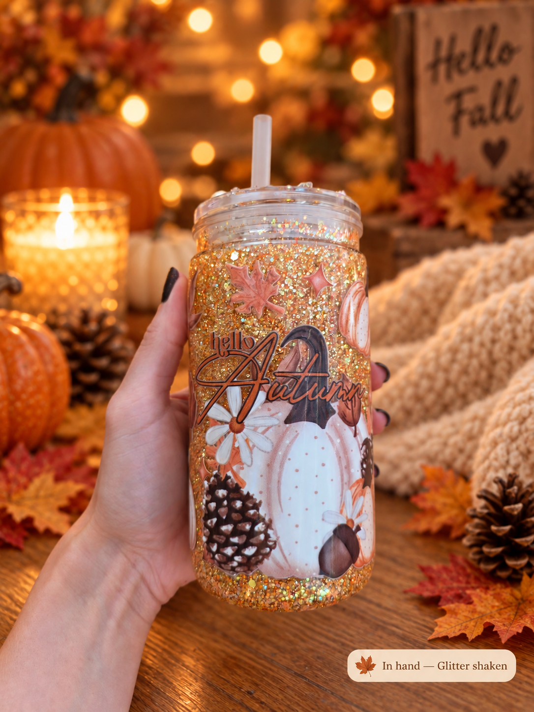 A hand holds a clear glitter tumbler with a straw, decorated with autumn-themed graphics including pumpkins, pinecones, acorns, and the words 'The Autumn'. The background is a cozy, warm fall scene with pumpkins, candles, a knitted blanket, and a wooden sign that reads 'Hello Fall'.