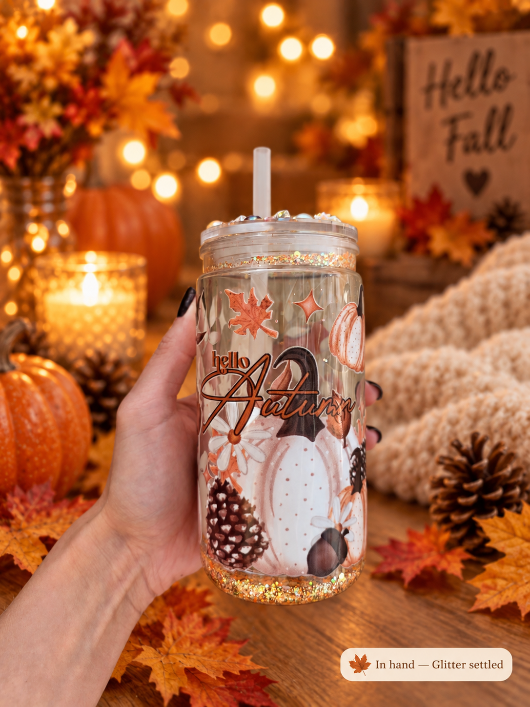 A hand holds a clear glitter tumbler with a white straw, decorated with autumn illustrations including pumpkins, pinecones, acorns, and the words 'hello nature'. The background is a cozy, warm fall scene with pumpkins, candles, autumn leaves, and a sign that reads 'hello fall'.