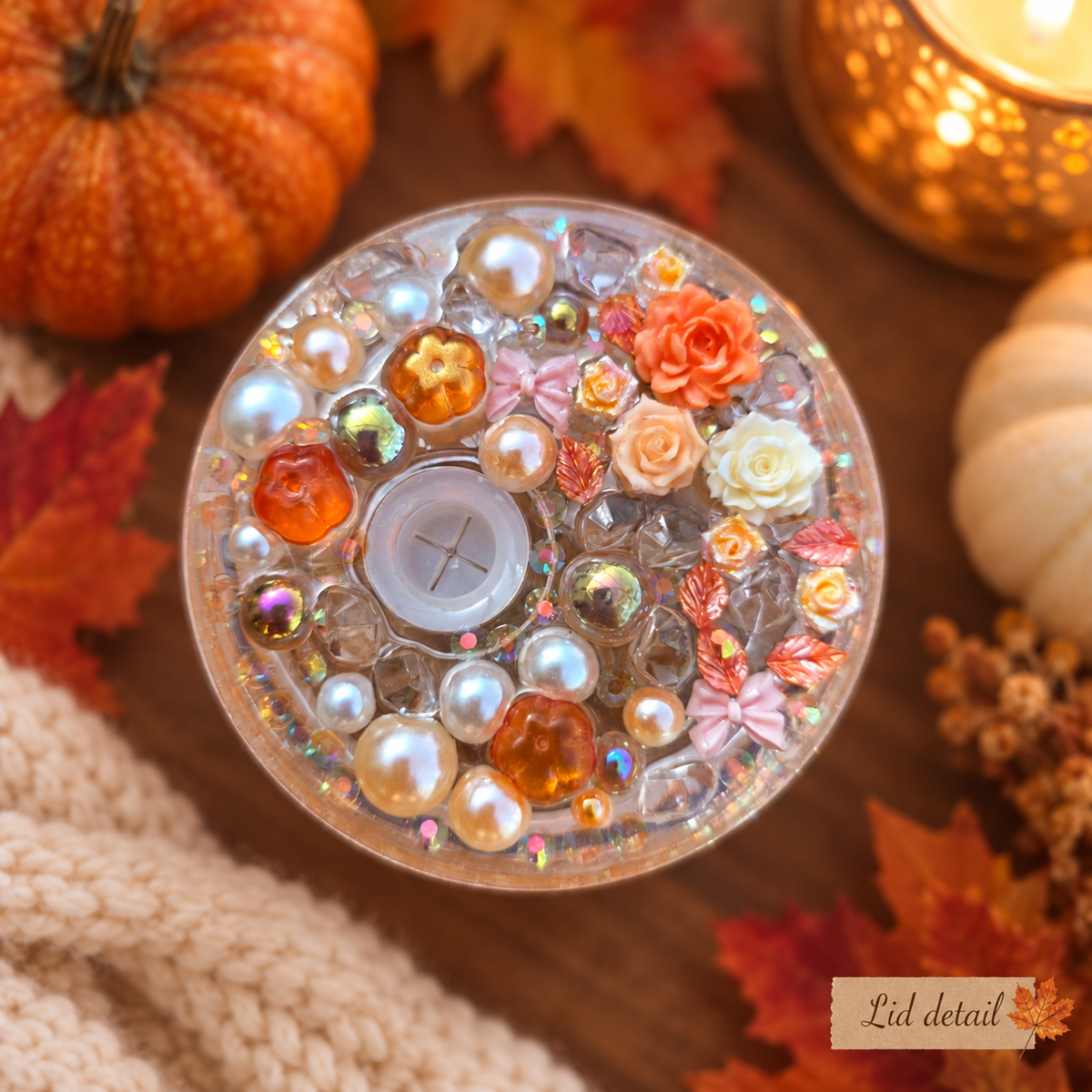 A round clear tumbler lid with decorative faux pearls, orange and cream flowers, and glitter, shown on a wooden surface with autumn leaves, pumpkins, and a candle. A small 'Lid detail' label is visible in the bottom right corner.