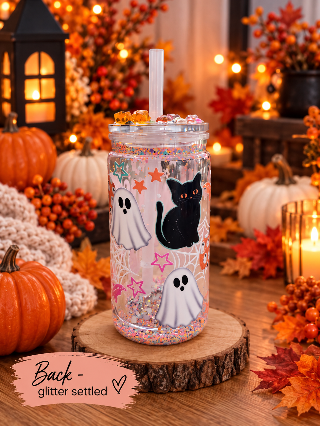 A clear glass mason jar tumbler with a white straw, decorated with a black cat, white ghosts, and pink glitter, displayed on a wooden slice coaster in a cozy, autumn-themed setting with pumpkins and candles.