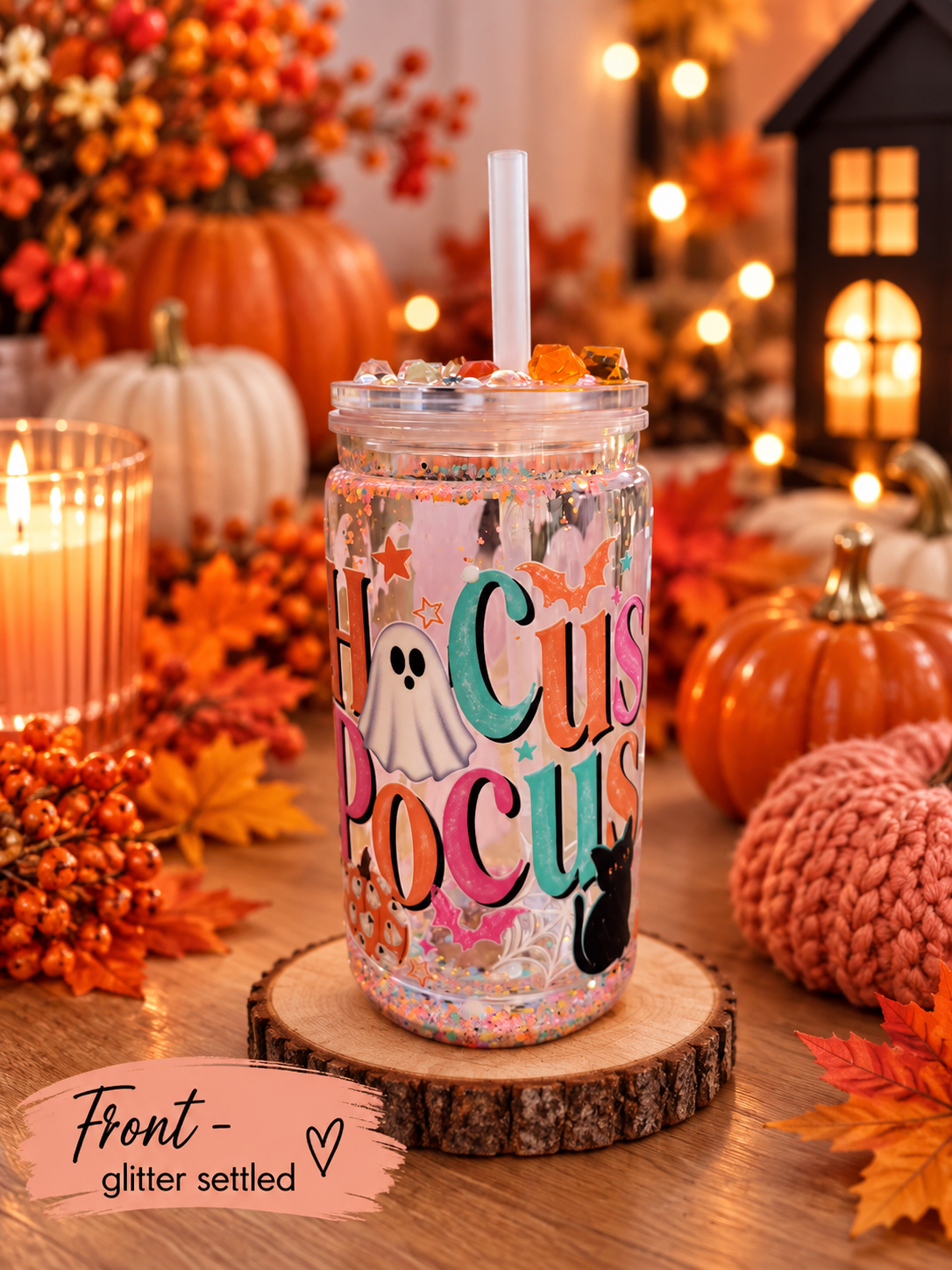 A clear glitter-filled mason jar tumbler with a white straw, decorated with 'Hocus Pocus' text, a ghost, and a black cat, displayed on a wooden slice coaster in a cozy autumn-themed setting with pumpkins, candles, and fall leaves.