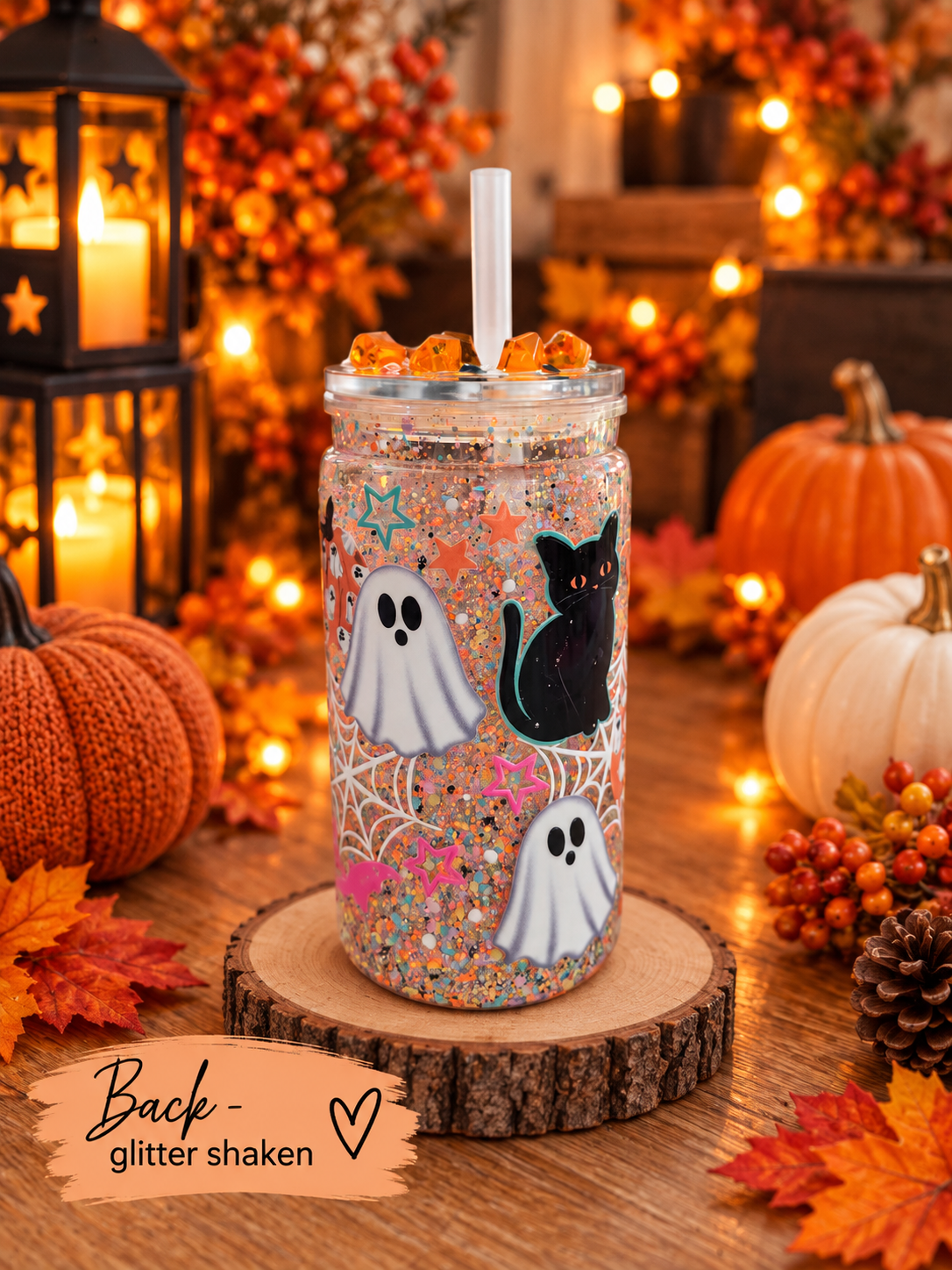 A Halloween-themed glitter shaker tumbler cup with a clear lid and straw, decorated with white ghosts, a black cat, and colorful glitter, shown on a wooden slice with autumn decorations in the background.