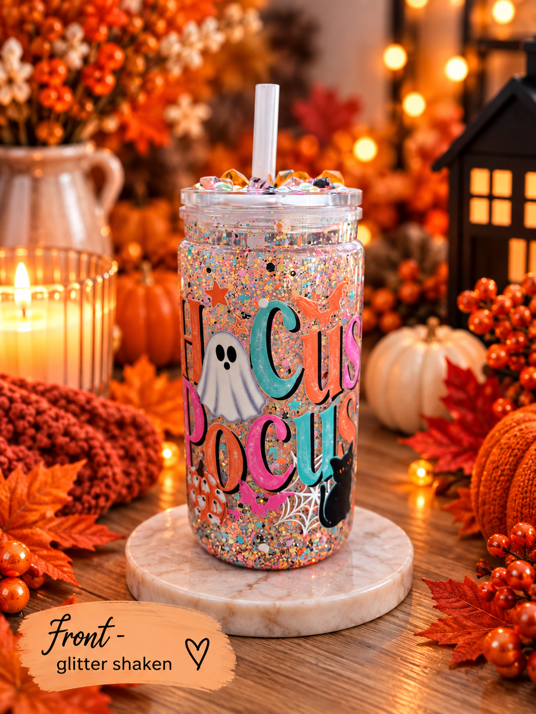 A clear plastic tumbler with a white straw sits on a round marble coaster in a fall-themed setting. The tumbler is decorated with multicolor glitter, a white ghost, black cat silhouette, and the words 'Hocus Pocus' in colorful lettering.