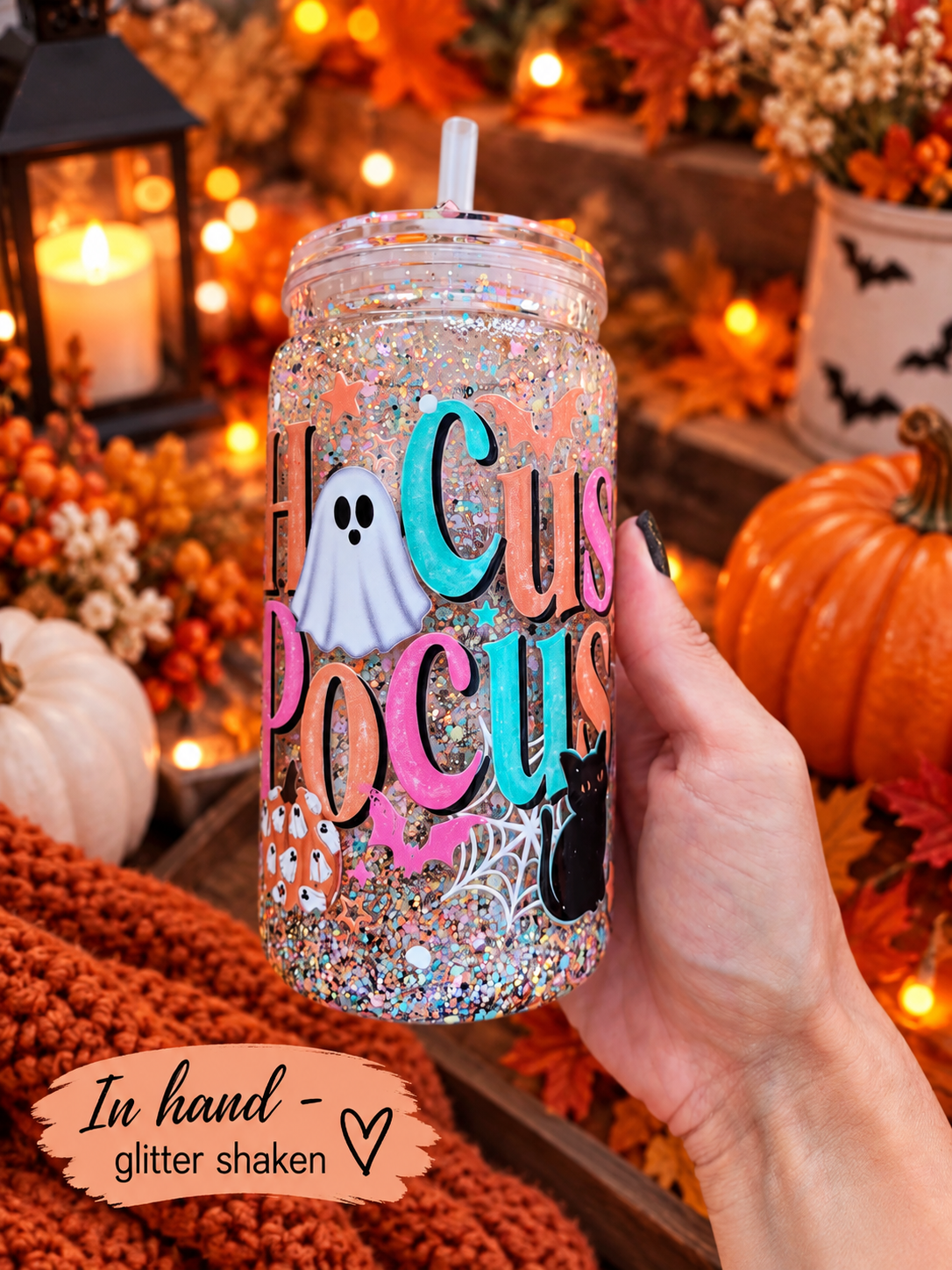 A hand holds a clear glitter tumbler with a straw, decorated with 'Hocus Pocus' text, a ghost, cat, spider, and pumpkin motifs. The background is a cozy autumnal setting with pumpkins, candles, and warm lights.