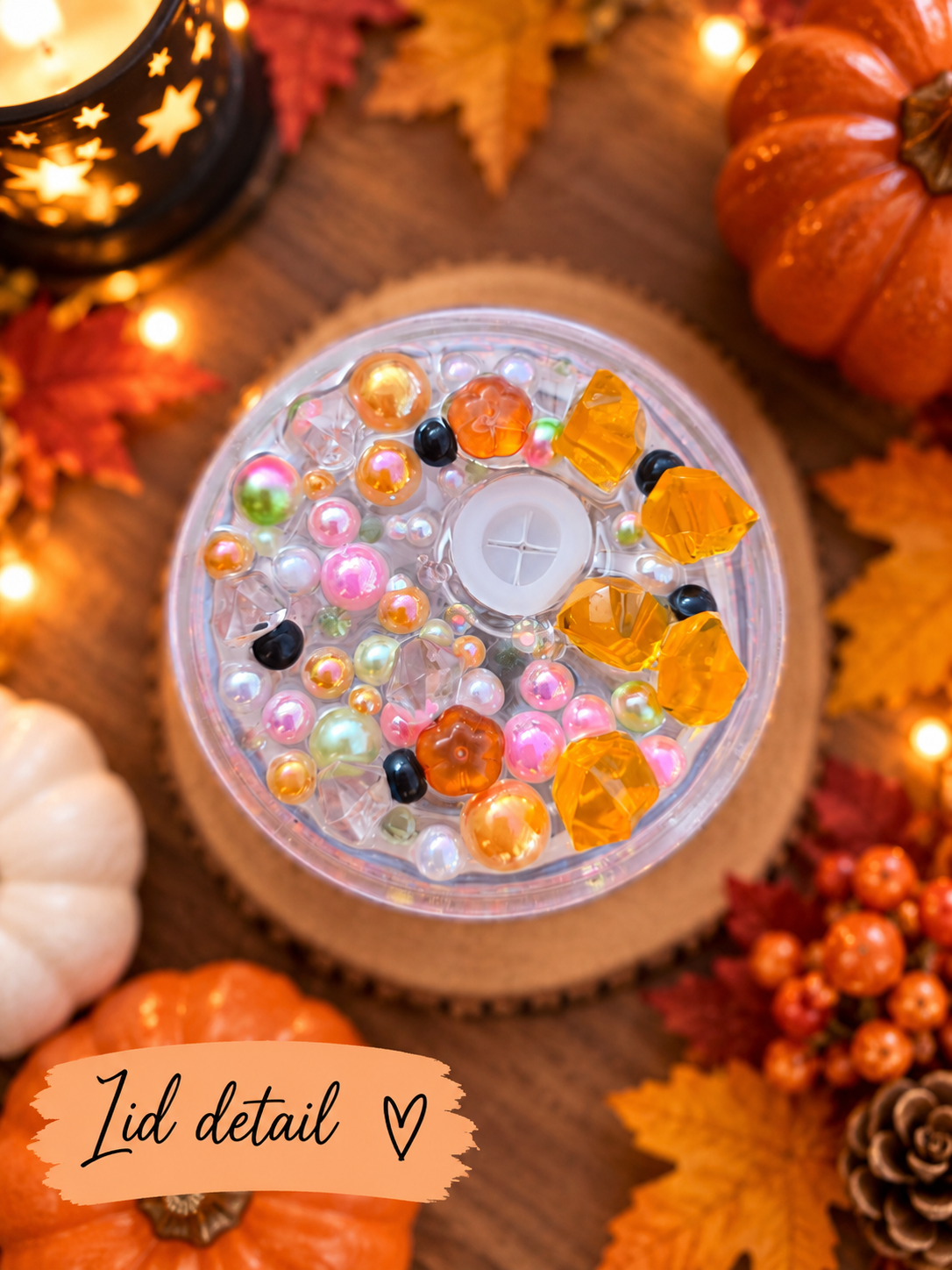 A clear round lid with colourful decorative beads and faceted orange gems, shown from above on a wooden surface with autumn leaves, pumpkins, and warm string lights around it. A label at the bottom reads 'Lid detail'.