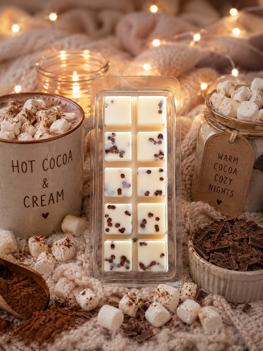 A clear plastic mold containing white wax melts with scattered dark chocolate pieces, styled on a cozy knit blanket with marshmallows, cocoa powder, a candle, and jars labeled 'HOT COCOA & CREAM' and 'WARM COCOA COZY NIGHTS'.