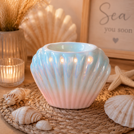 A seashell-shaped ceramic wax and oil holder with a glossy iridescent finish, glowing with warm light from built-in candles. It is styled on a woven placemat with seashells, a lit glass candle holder, dried grass, and a framed sign in the background.