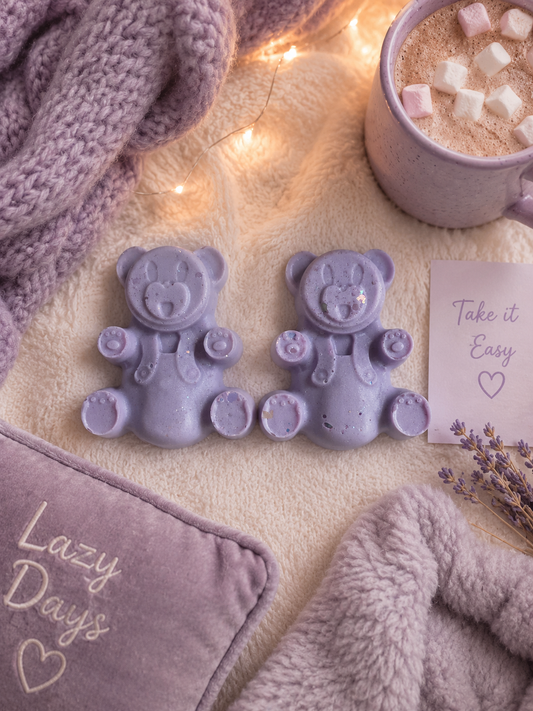 Two lavender purple bear-shaped wax melts displayed on a cozy beige blanket with a knit purple scarf, a mug of hot chocolate, a lavender pillow, and a card that reads 'Take it Easy'.