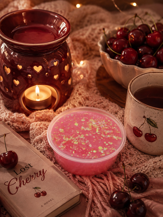 A styled cozy scene with a pink cherry-gel wax melt in a clear container, a lit candle in a heart-lit amber holder, cherries in bowls, a mug with cherry illustrations, and a book titled "Lost Cherry.".