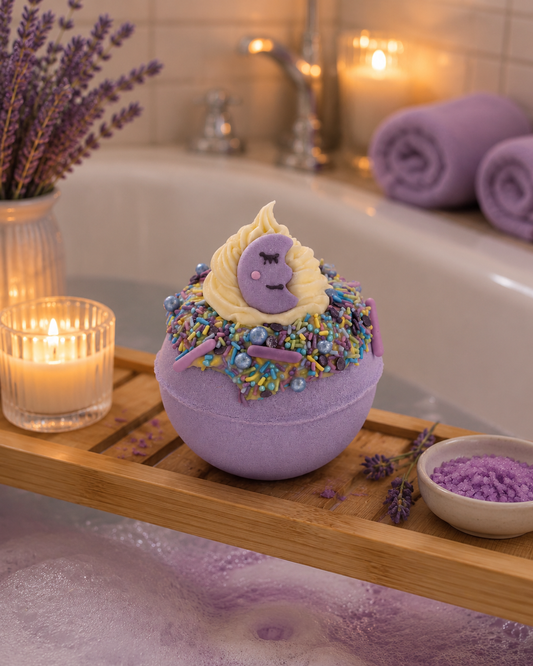 A lavender bath bomb sits on a wooden bath tray in a bubble bath, decorated with a cream swirl, purple flower, and colorful sprinkles. Lavender sprigs, a small bowl of bath salts, and rolled towels are arranged around it in a cozy bathroom setting.