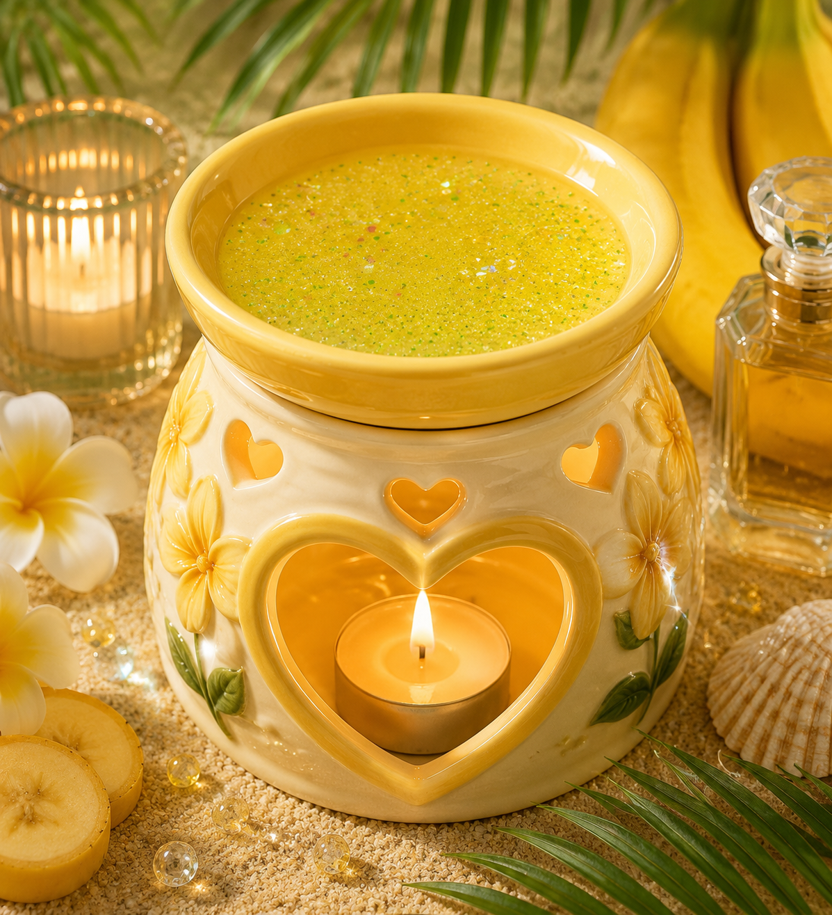 A ceramic candle warmer with a yellow lid and heart-shaped cutout base, lit with a small tealight candle with gel wax inside. The warmer is styled with tropical props including plumeria flowers, sliced bananas, a glass jar candle, a perfume bottle, and green palm leaves on a sandy surface.