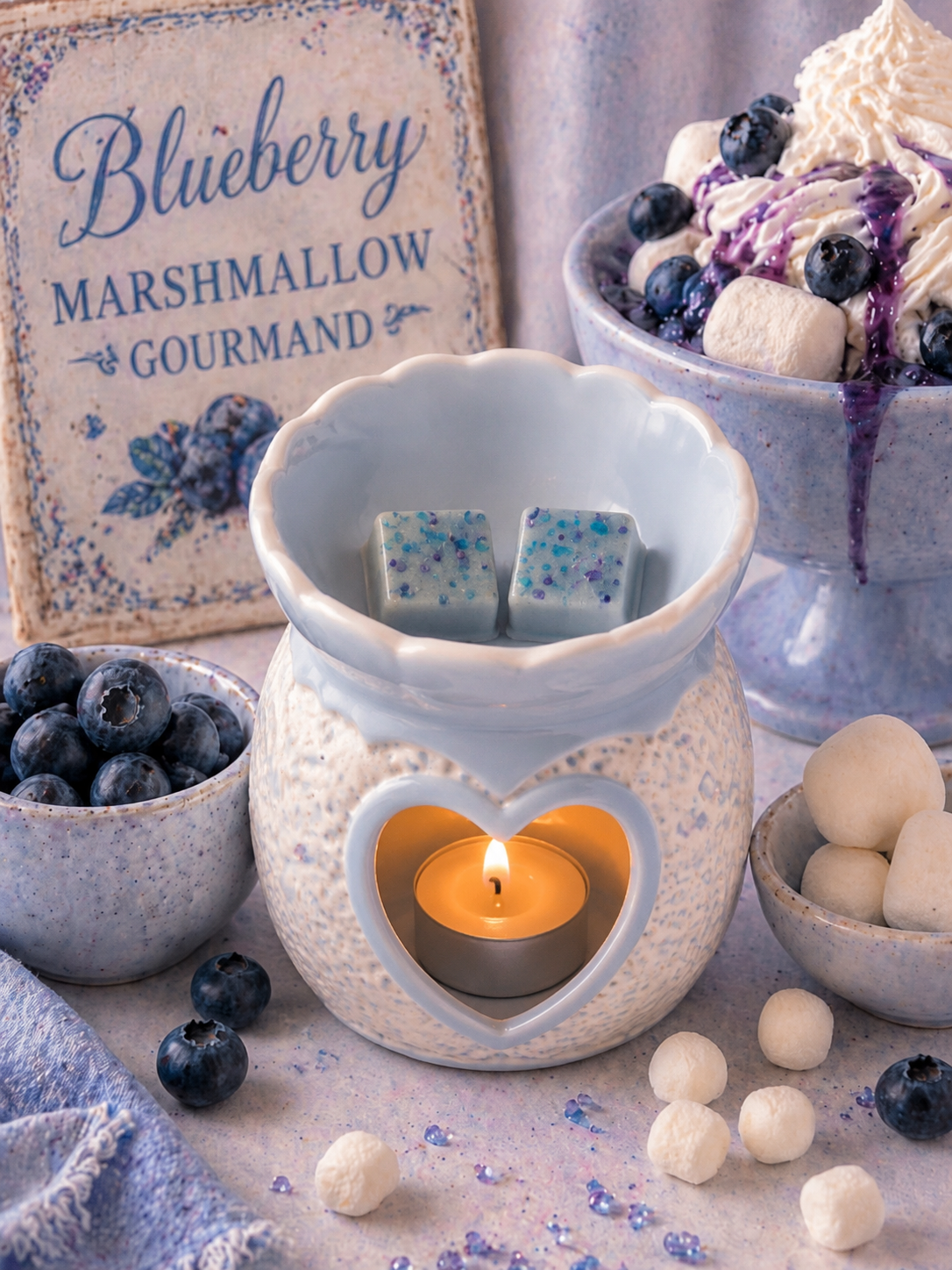 A ceramic blueberry-themed candle warmer with a lit tealight candle inside a heart-shaped cutout, topped with two blueberry-infused wax cubes. The scene is styled with blueberries, marshmallows, and a sign reading 'Blueberry Marshmallow Gourmand'.