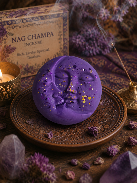 A purple wax melt shaped like a crescent moon with a face and scattered gold leaf flakes, displayed on an ornate bronze tray. A box labeled 'Nag Champa Incense' and a burning incense burner are visible in a styled setting with dried lavender, crystals, and a candle.