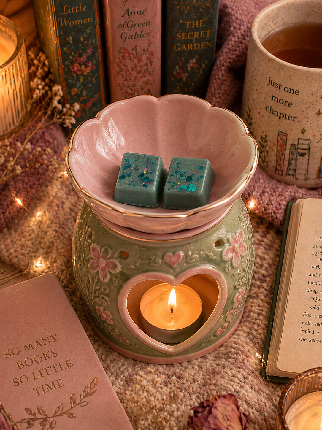 A ceramic heart-shaped candle warmer with a pink floral top and a mint green body, holding a lit teal candle and two teal wax cubes with glitter. The warmer is styled on a knit blanket with books, tea, and fairy lights in the background.