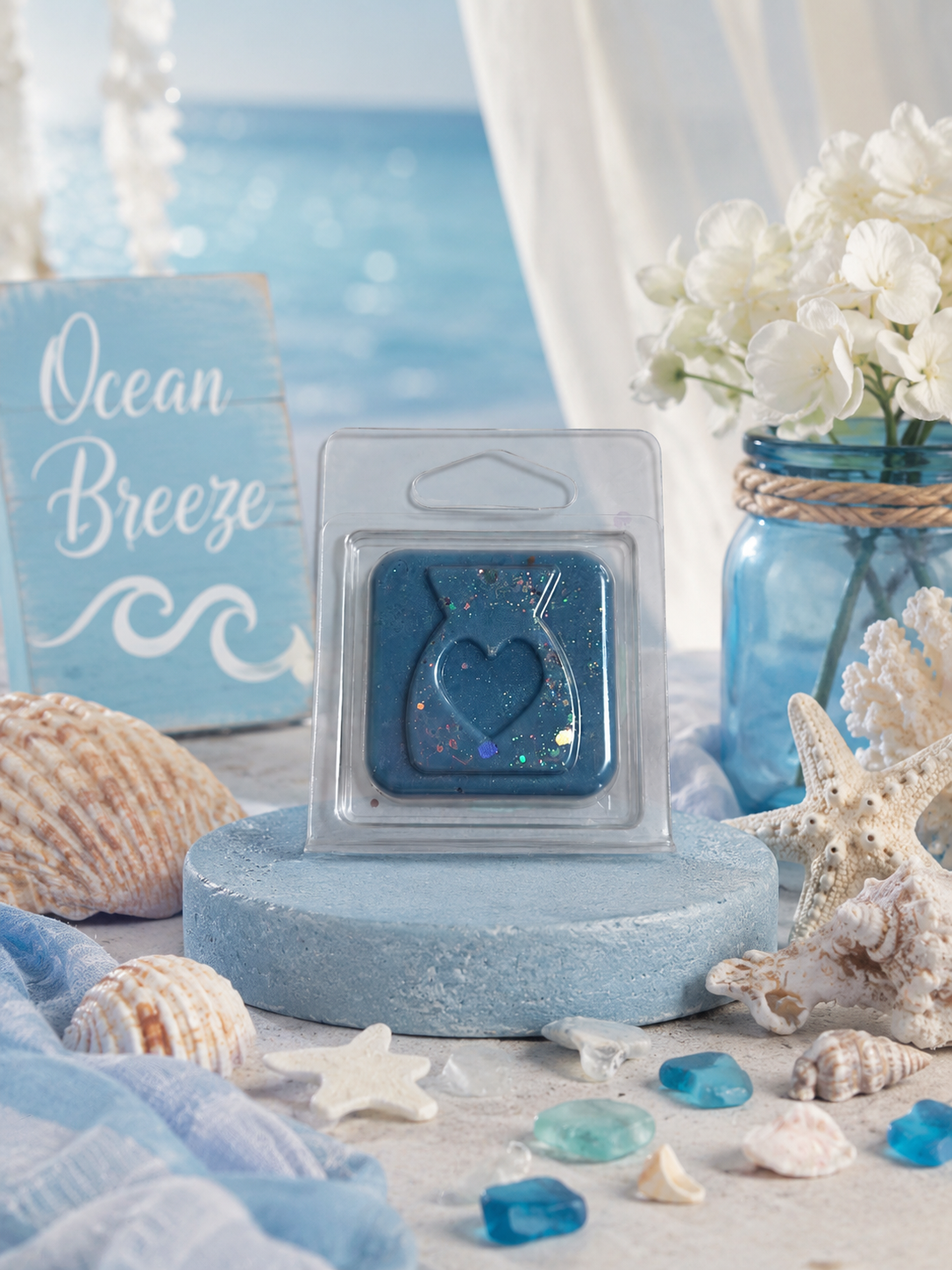 Blue wax melt with heart design in packaging on a beach-themed surface with shells and flowers.