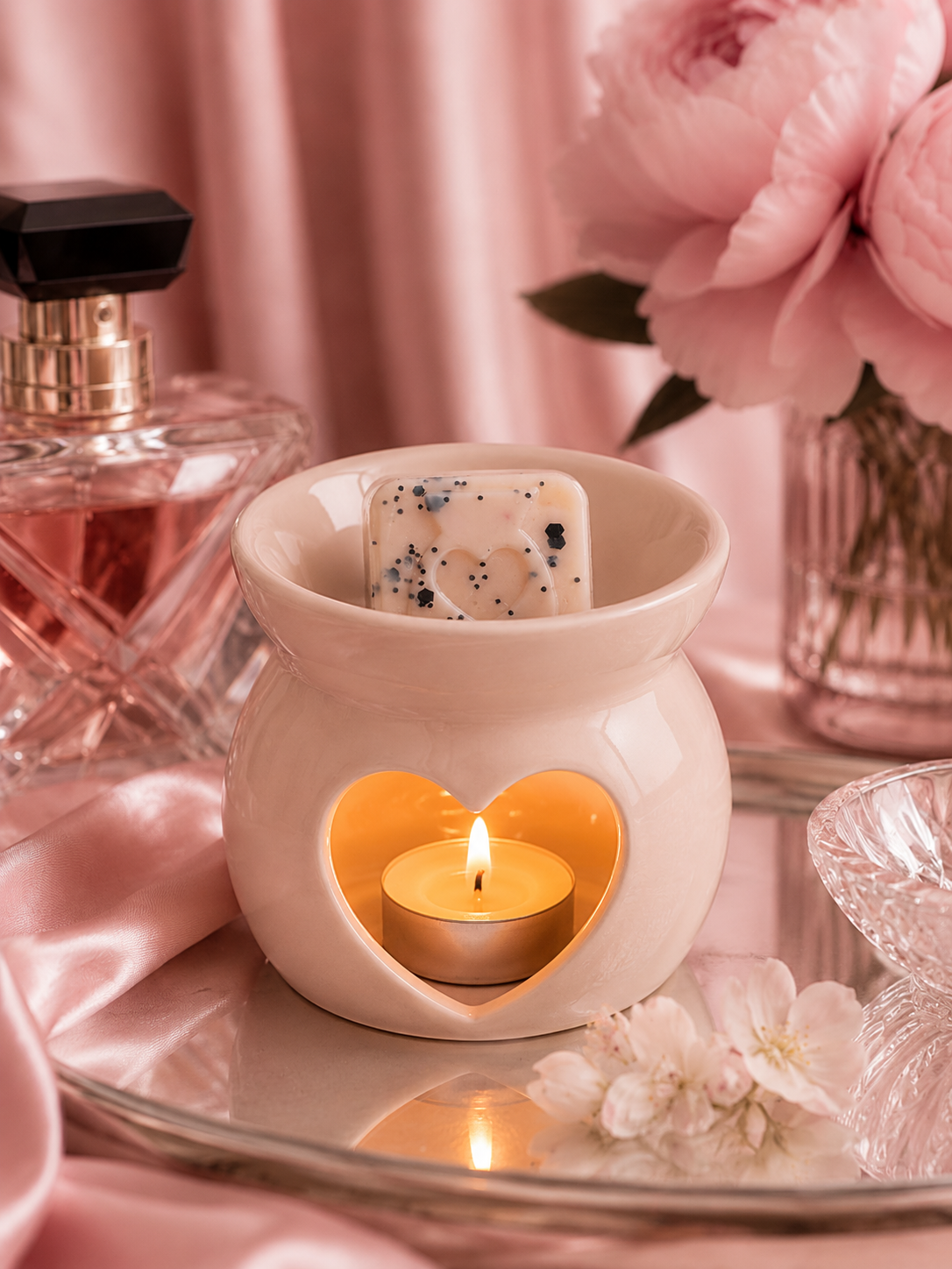 A pink ceramic heart-shaped candle warmer holding a lit tealight candle, styled on a mirrored tray with pink satin fabric, flowers, and a perfume bottle in the background.