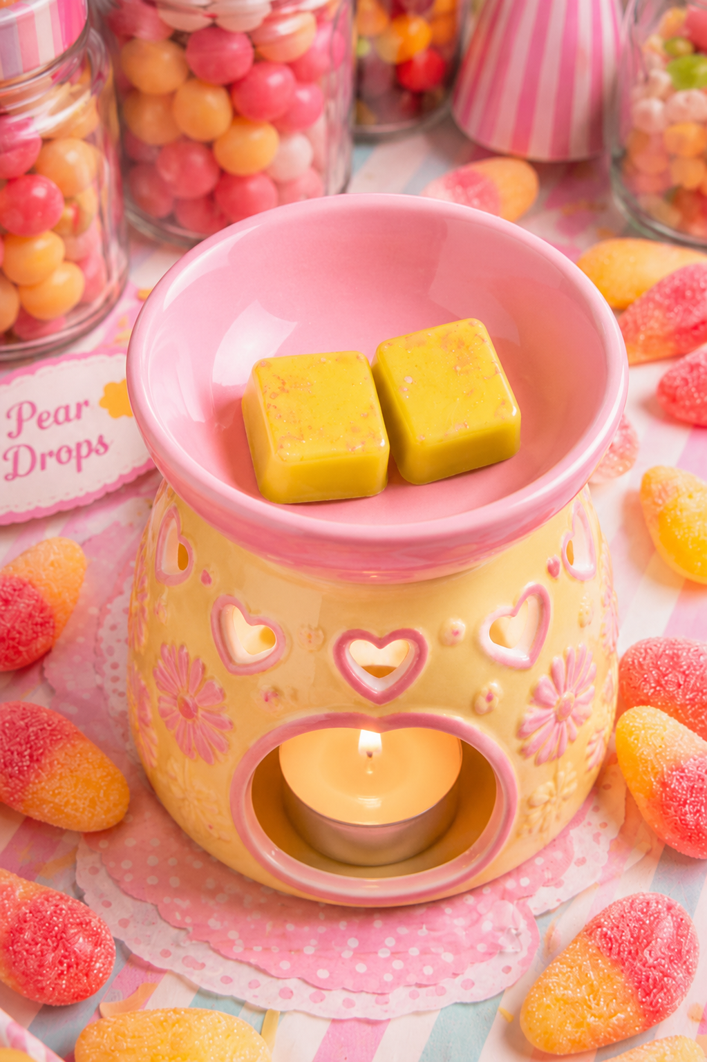 A pink and yellow ceramic wax warmer with heart and flower details, holding two yellow wax cubes and a lit tealight candle, surrounded by candy on a pastel striped surface.