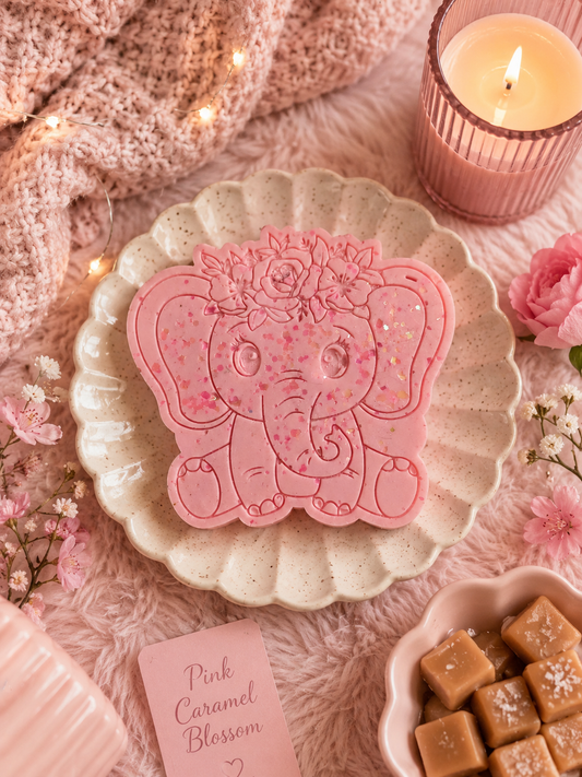 A pink elephant-shaped decorative item or candle sits on a scalloped cream plate, styled with a knitted pink scarf, a lit candle, flowers, and caramel candies on a soft pink surface.