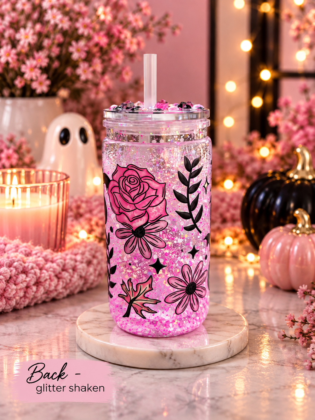 A pink glitter shaker tumbler with black floral illustrations sits on a marble coaster in a cozy, autumn-themed setting with pumpkins, a candle, and fairy lights.