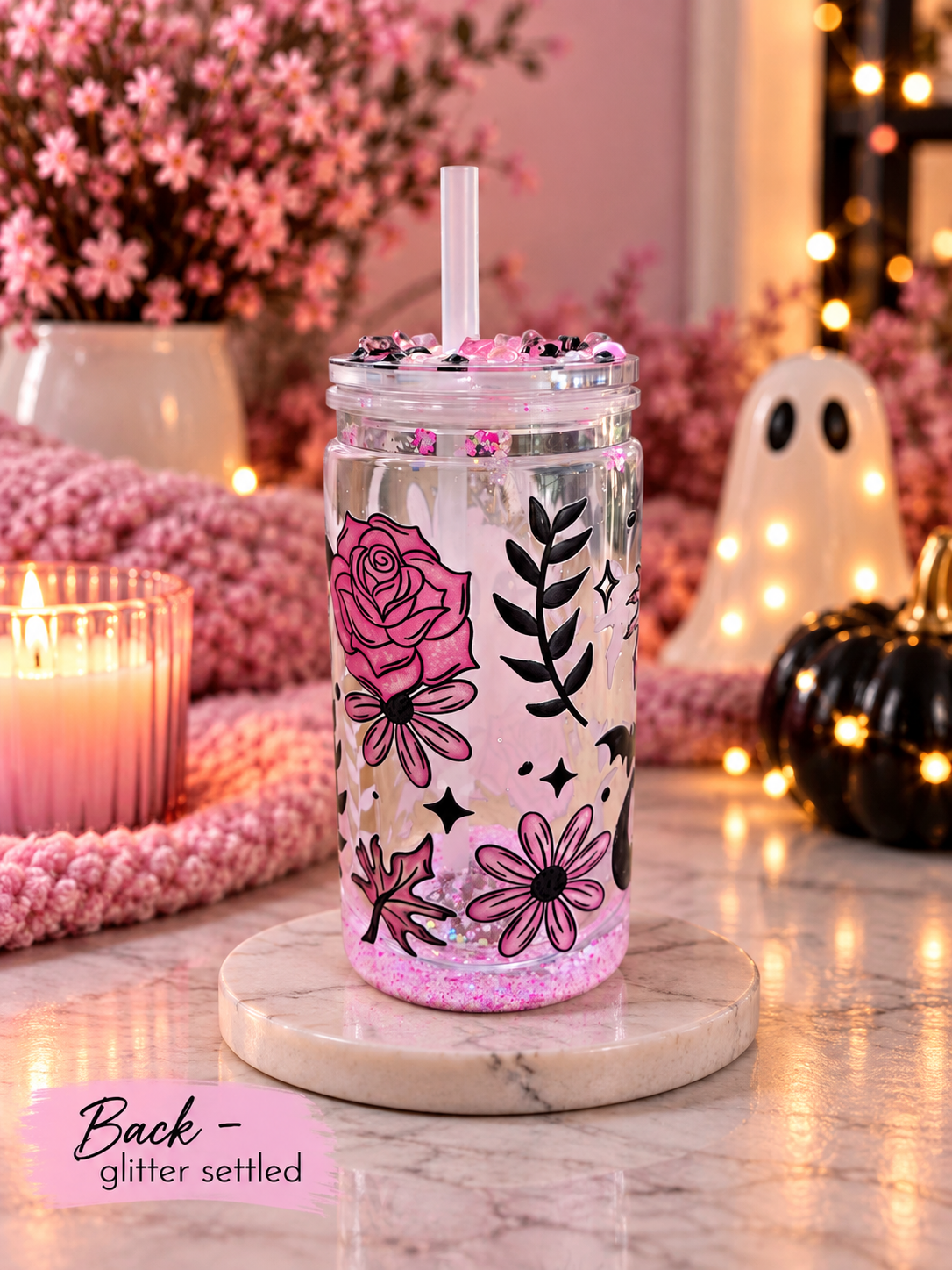 A clear tumbler with a white straw, decorated with pink and black floral illustrations and glitter, sitting on a marble coaster in a cozy, Halloween-themed setting with candles, pumpkins, and fairy lights.
