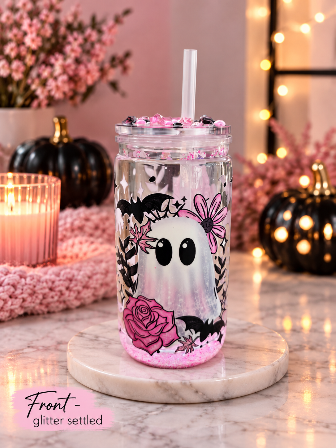 A clear tumbler with a white ghost, pink roses, and black bats printed on the front, filled with pink glitter and a clear straw, set on a marble coaster in a cozy fall-themed setting.