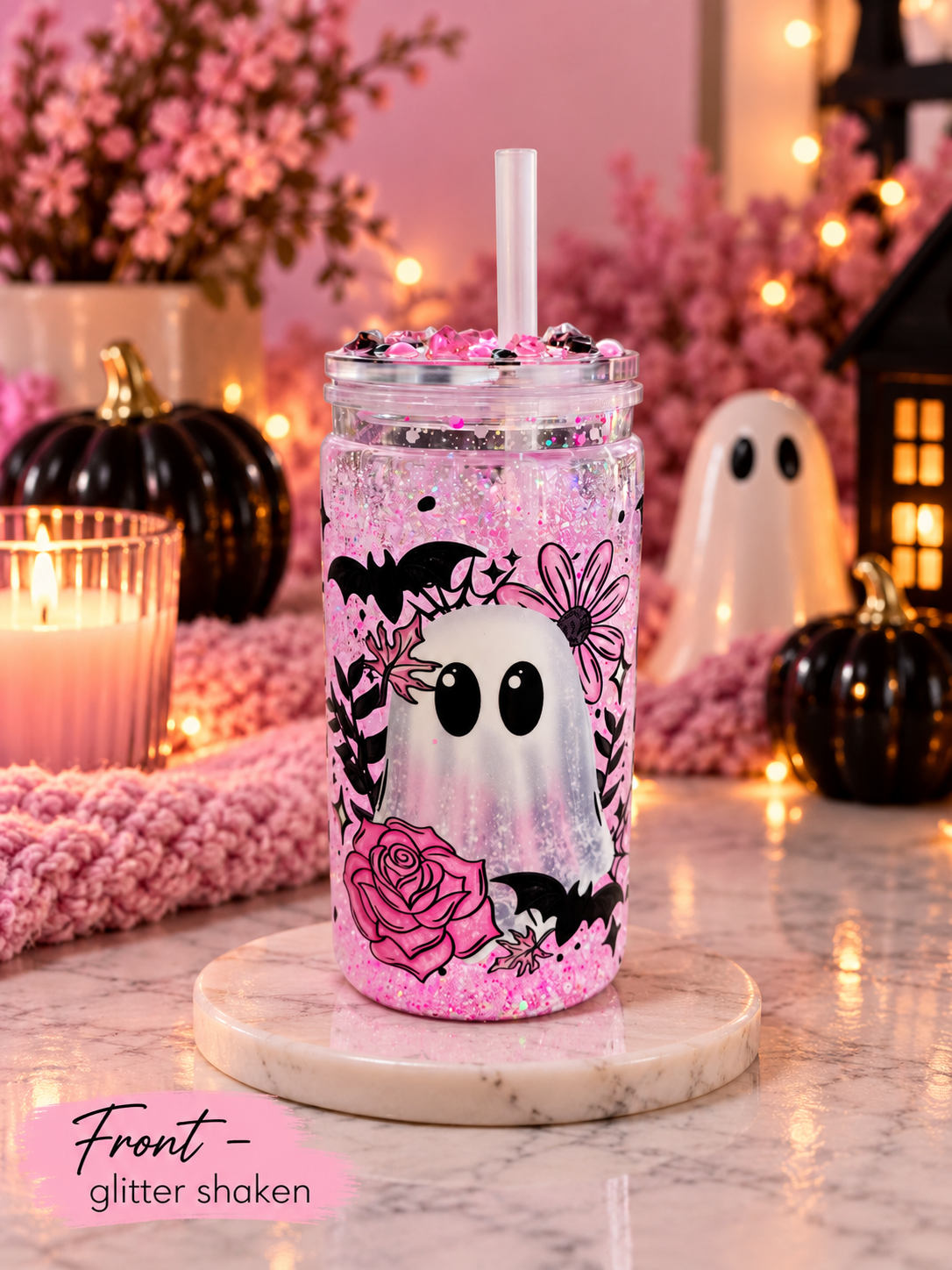 A clear plastic tumbler with a straw, filled with pink glitter and topped with pink sprinkles, decorated with a black and white ghost, pink rose, and bat design. The tumbler is displayed on a marble coaster in a cozy, Halloween-themed setting with pink yarn, black pumpkins, and fairy lights.