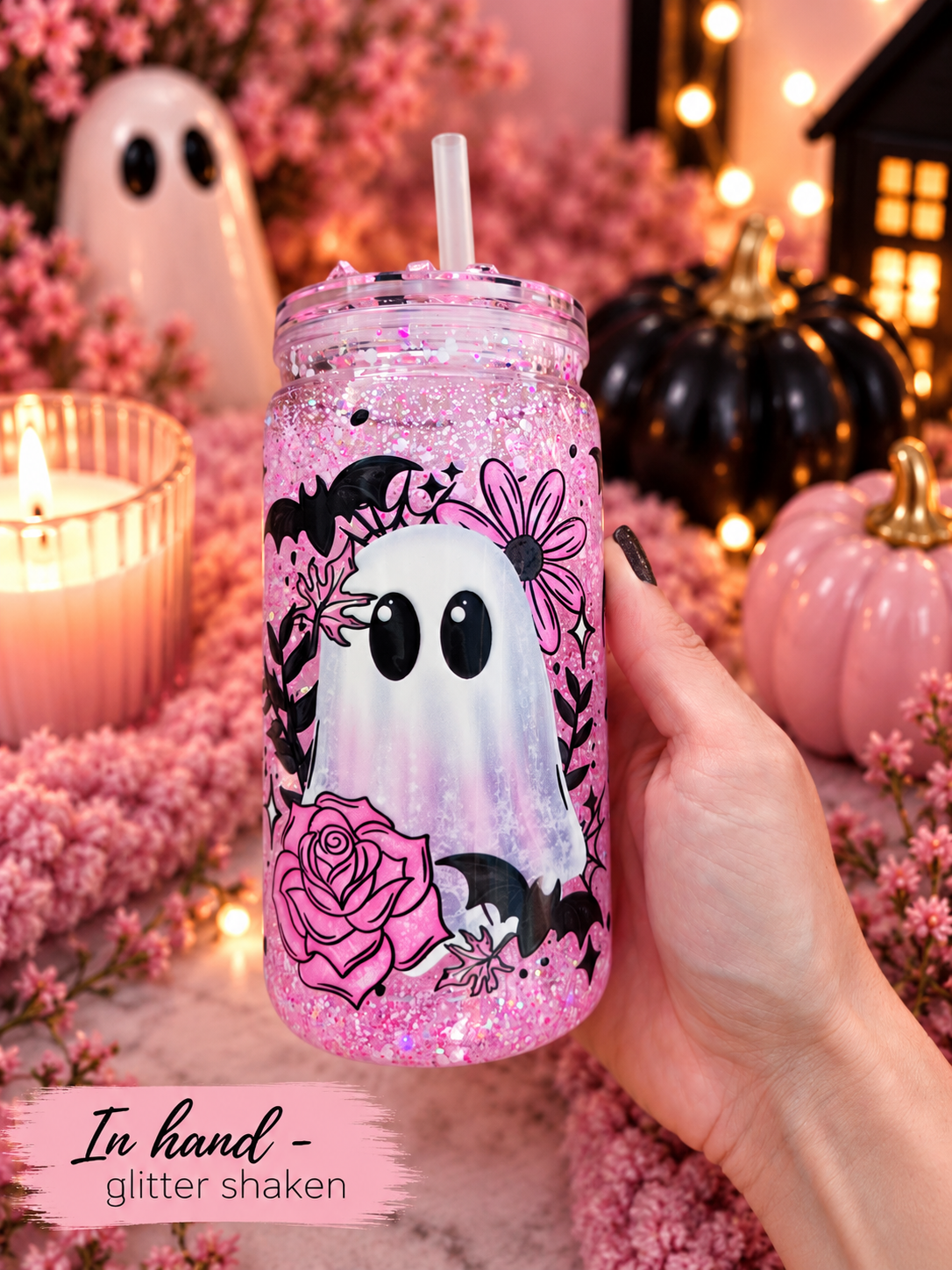 A hand holds a pink glitter tumbler with a straw, decorated with a Halloween ghost design, in a cozy, festive setting with pumpkins, candles, and pink flowers.