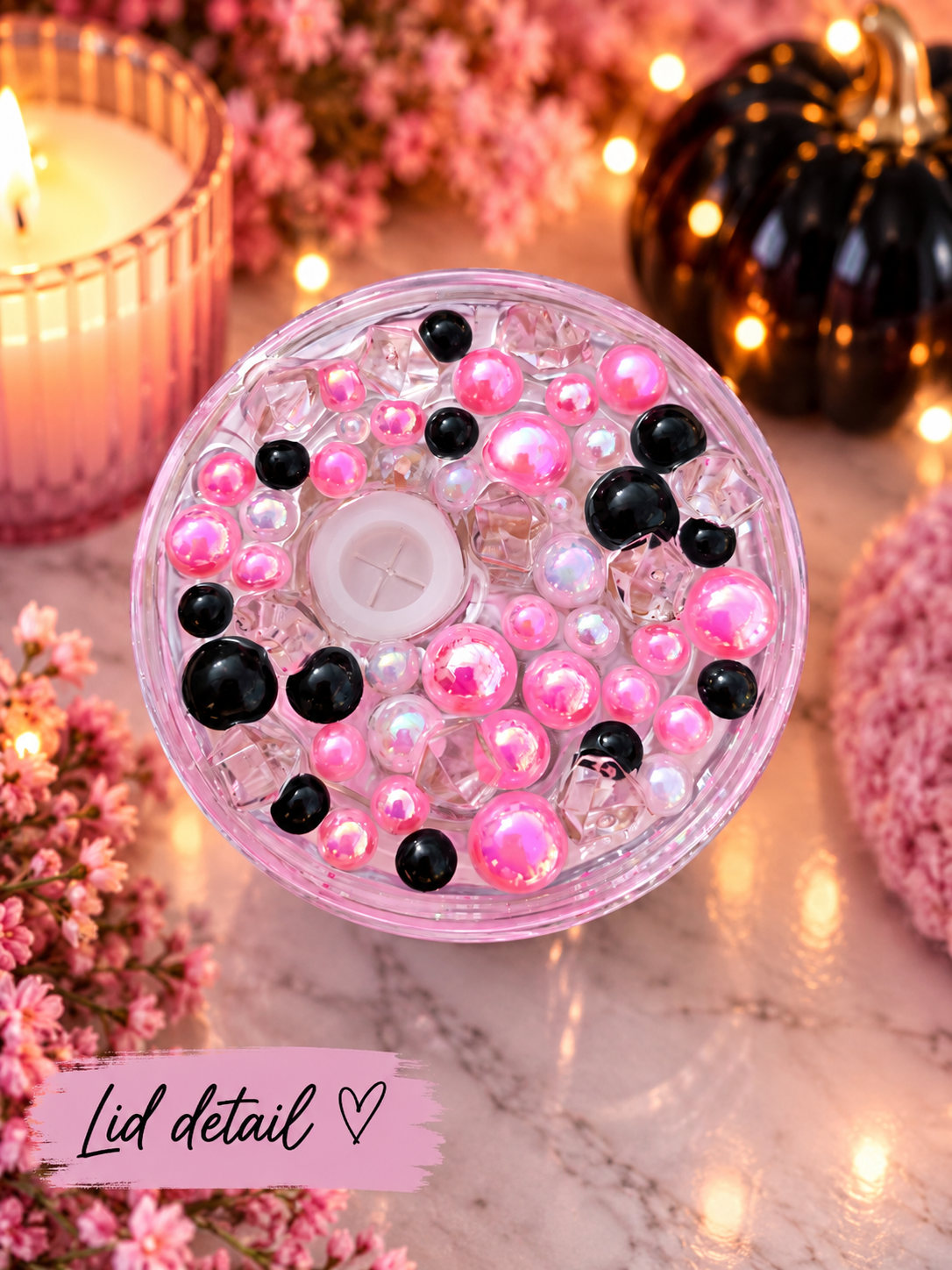 A round lid filled with pink and black decorative beads, pearls, and a white button, shown on a marble surface with candles, flowers, and warm string lights around it. A pink label at the bottom reads 'Lid detail'.