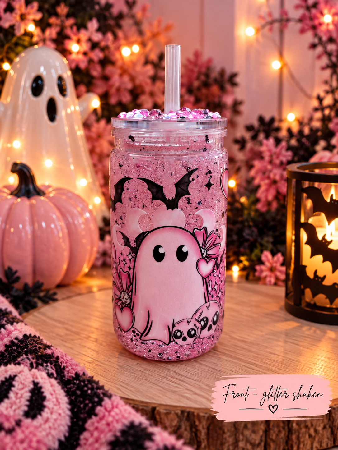 A pink glitter-filled mason jar tumbler with a clear straw, decorated with a cute cartoon ghost and cat illustration, set on a wooden table in a cozy Halloween-themed setting with a glowing ghost decoration, a pink pumpkin, and a candle.