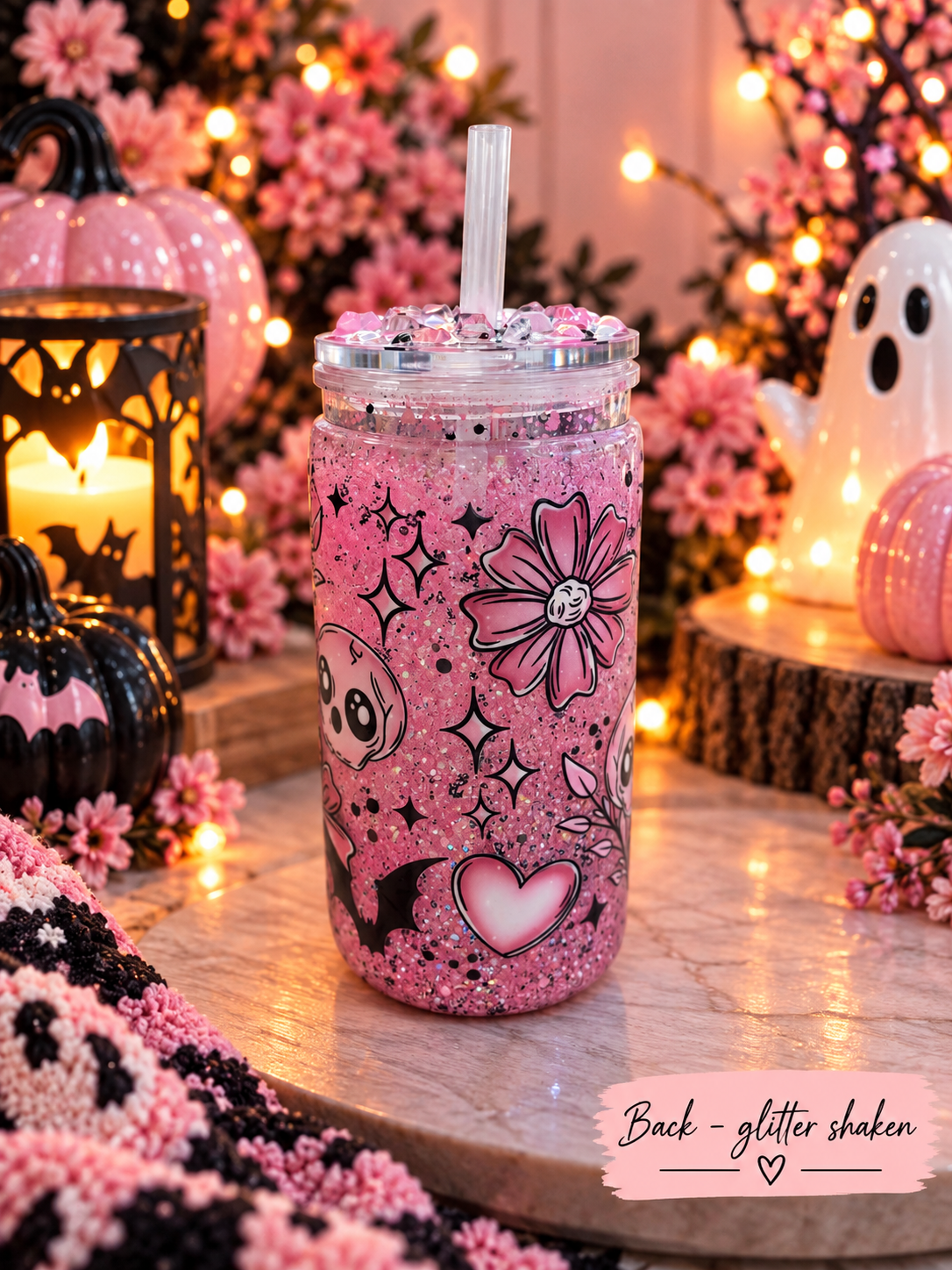 A pink glitter-shaken drink in a clear mason jar with a straw, decorated with black doodles, flowers, stars, and a heart, set on a tabletop with Halloween decorations in the background.