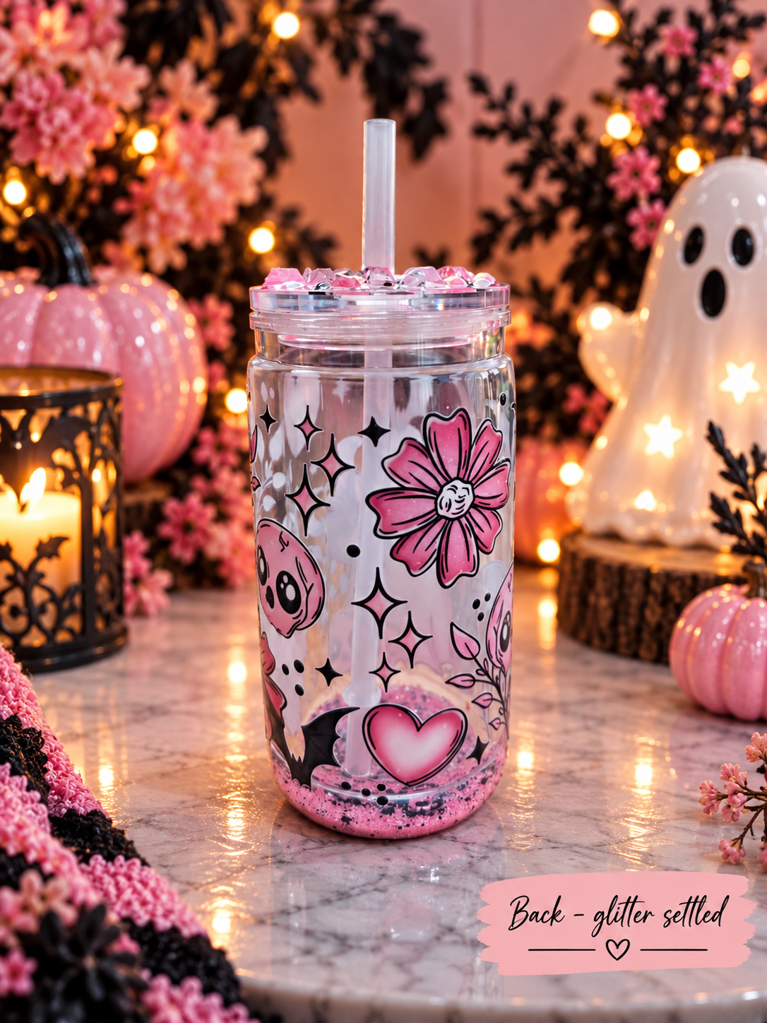 A clear plastic tumbler with a straw, decorated with pink glitter and a Halloween-themed print featuring a skull, flower, and heart. The tumbler is set on a marble surface surrounded by cozy fall decorations, including a ghost, pumpkins, and candles.