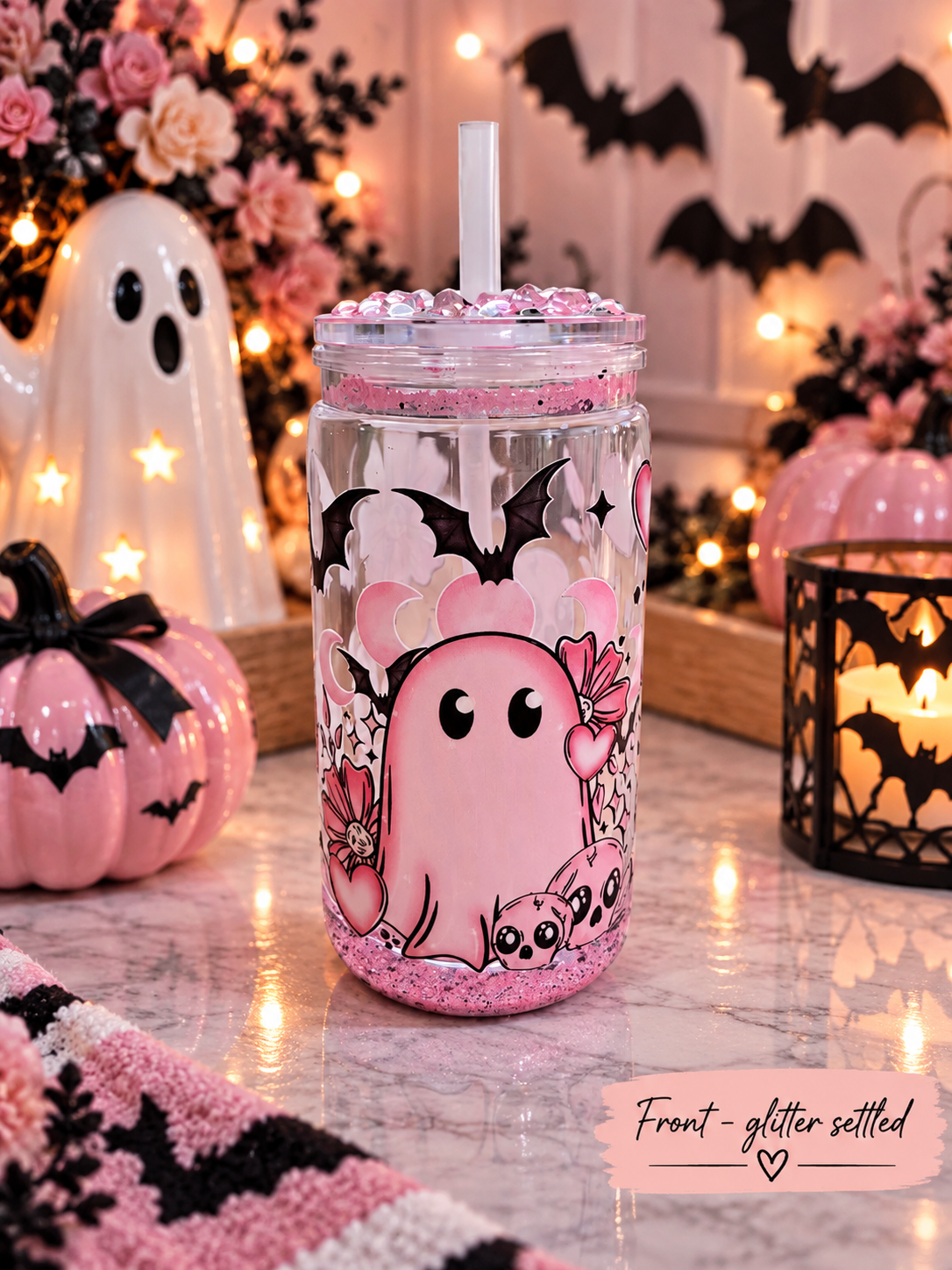 A clear tumbler with a pink glitter base and a white straw, decorated with a cute pink ghost, black bats, and floral designs. The tumbler is set on a marble table surrounded by Halloween decorations, including a ghost figurine, a pink pumpkin, and a candle holder.