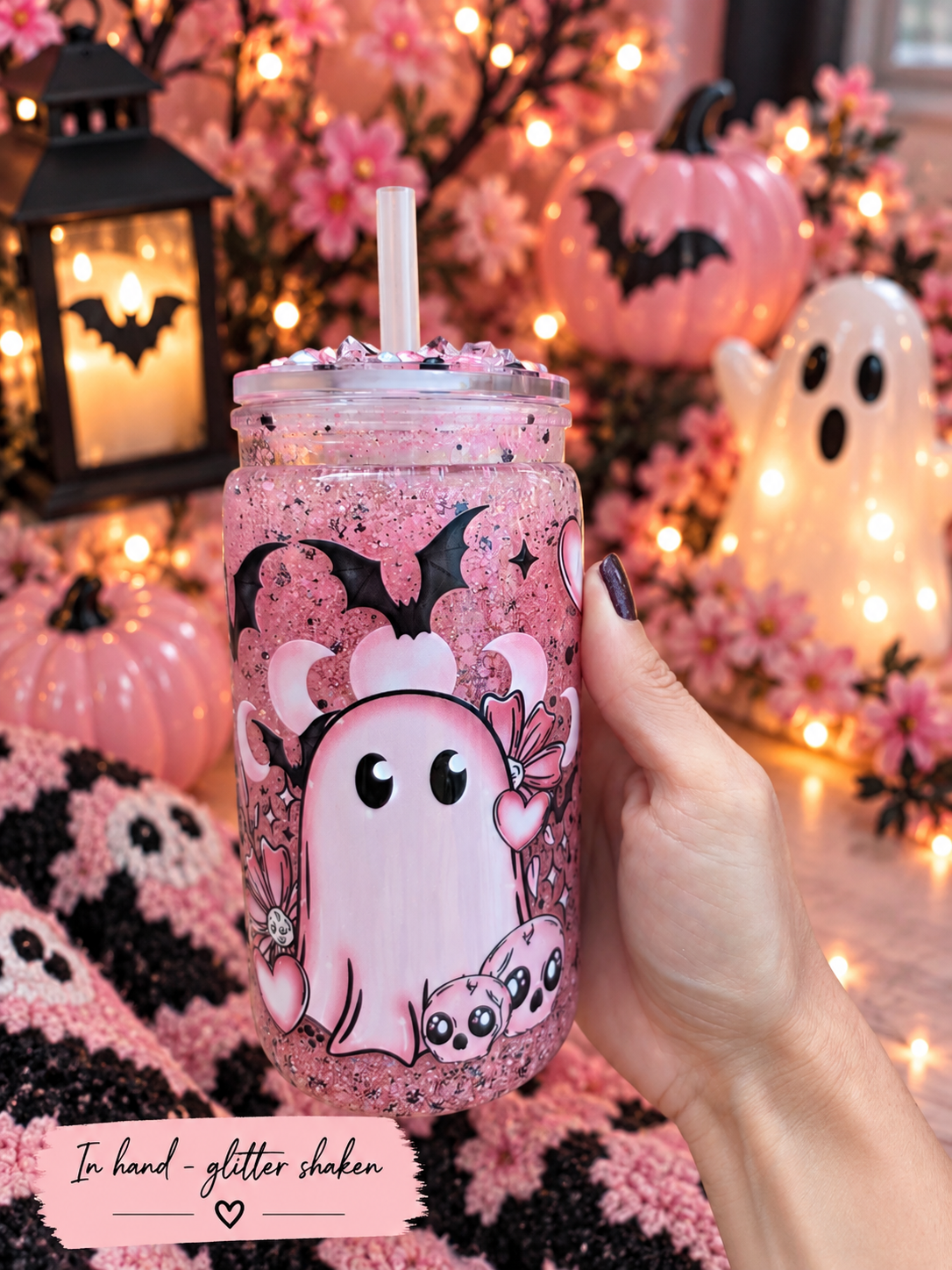A hand holds a pink glitter tumbler with a straw, decorated with a cute cartoon ghost and cat design. The background is a cozy, festive Halloween scene with glowing pumpkins, a lantern, and a ghost decoration.
