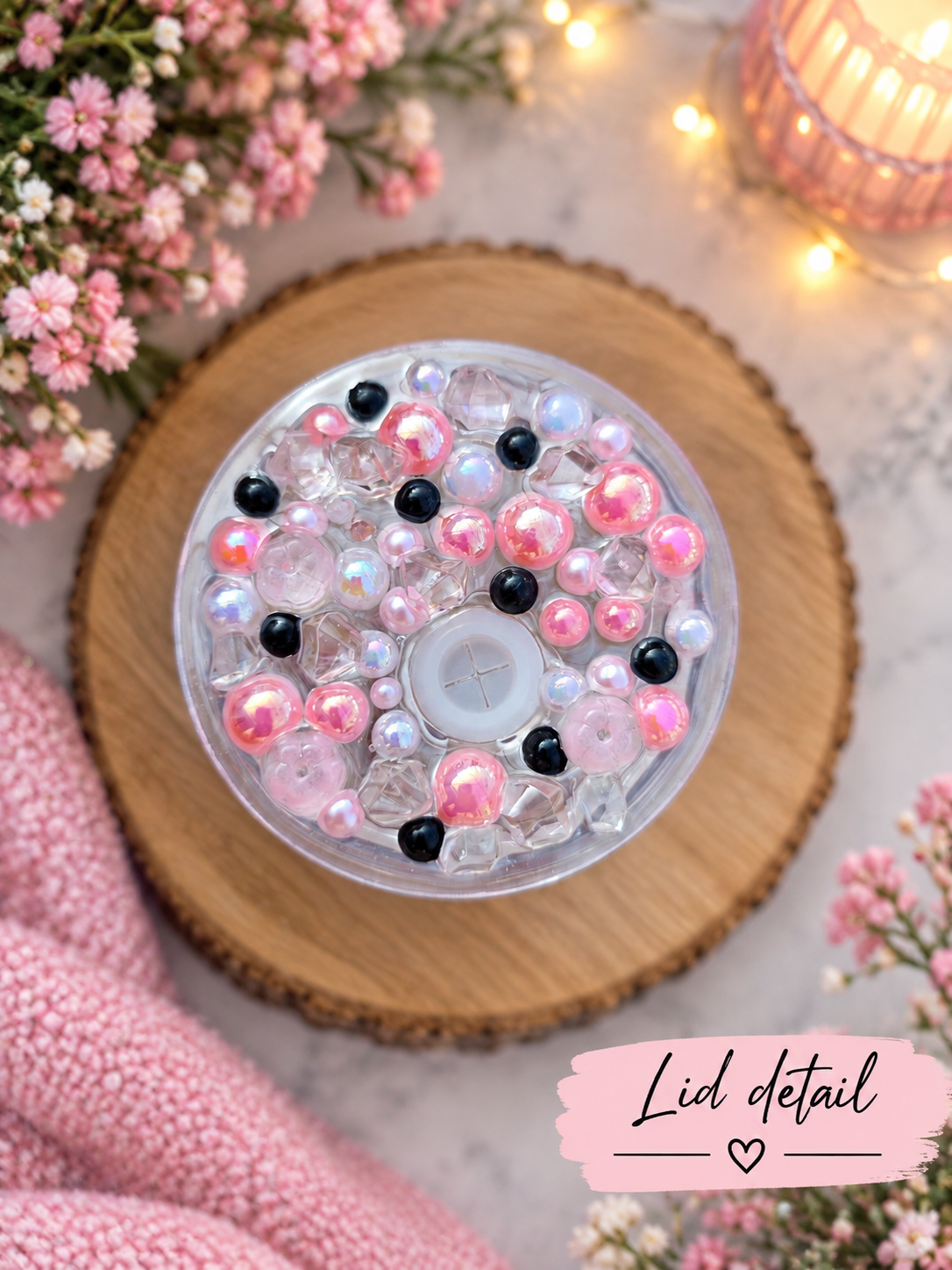 Top-down view of a round clear plastic tumbler lid with pink, black, and iridescent beads, placed on a round wooden slice with pink flowers and fairy lights around it. A pink label in the corner reads 'Lid detail'.
