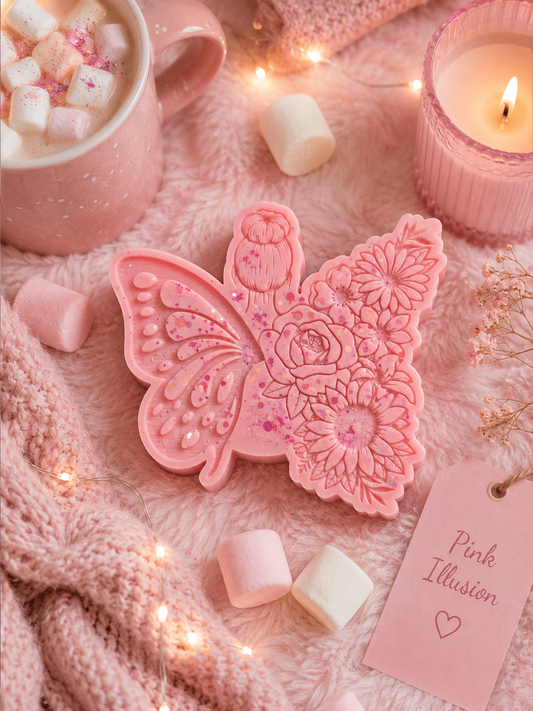 A pink butterfly-shaped decorative item with floral embossed details, styled on a soft pink blanket with marshmallows, a pink mug of hot chocolate, a lit candle, and a tag reading 'Pink Illusion'.