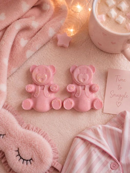 Two pink teddy bear-shaped wax melts on a soft pink blanket next to a pink sleep mask, pajamas, a mug of hot chocolate, and a card that reads 'Time to Snuggle'.