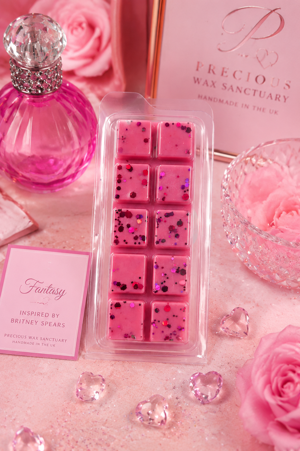 A pink wax melt pack in a clear plastic clamshell container, styled with pink roses, a pink perfume bottle, and branded cards on a pink surface. The visible text includes 'Precious Wax Sanctuary' and 'Fantasy Inspired by Britney Spears'.