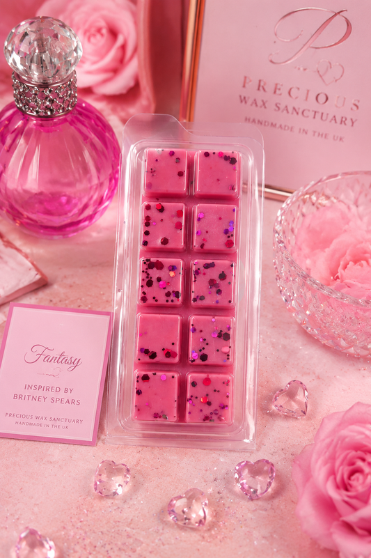 A pink wax melt pack in a clear plastic clamshell container, styled with pink roses, a pink perfume bottle, and branded cards on a pink surface. The visible text includes 'Precious Wax Sanctuary' and 'Fantasy Inspired by Britney Spears'.