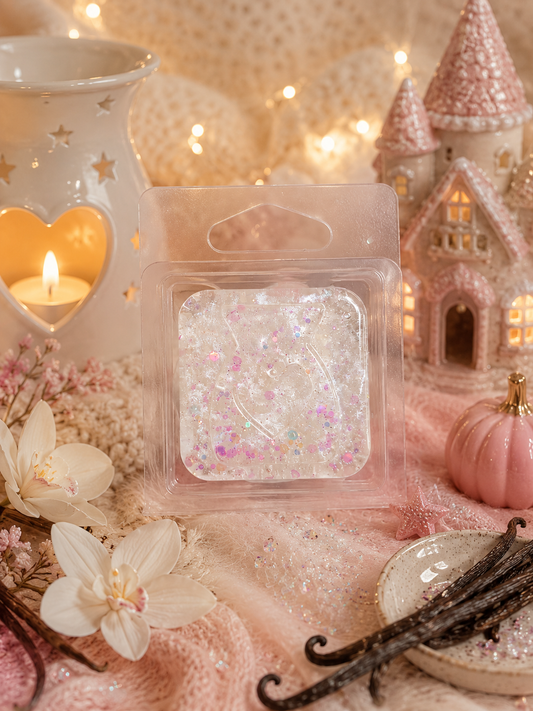 A clear plastic wax melt package containing a square gel white wax melt filled with pink and iridescent glitter, styled on a soft pink fabric with a candle holder, flowers, vanilla beans, and a decorative castle.
