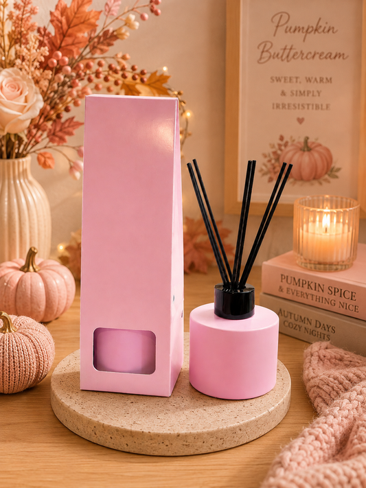 Pink reed diffuser bottle with black reeds next to its tall pink box, styled on a round coaster with fall decor, a candle, and a cozy knit blanket.