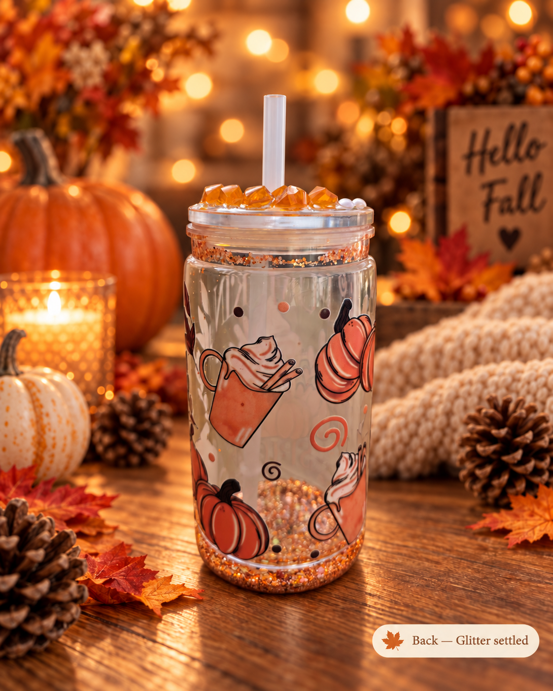 A clear mason jar tumbler with a white straw, decorated with orange pumpkins, hot cocoa illustrations, and glitter settled at the bottom, displayed on a wooden table in a cozy autumn setting with pumpkins, candles, and fall leaves.
