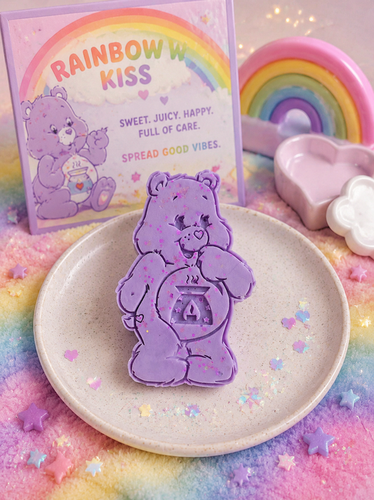 A pastel lavender Care Bear-shaped wax melt displayed on a speckled cream plate, with a matching 'Rainbow Kiss' branded card in the background and rainbow-themed decorative props.